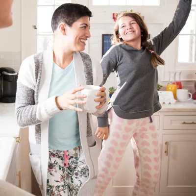 A woman dancing in the kitchen with her daughters and drinking coffee.