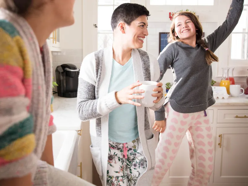 A woman dancing in the kitchen with her daughters and drinking coffee.